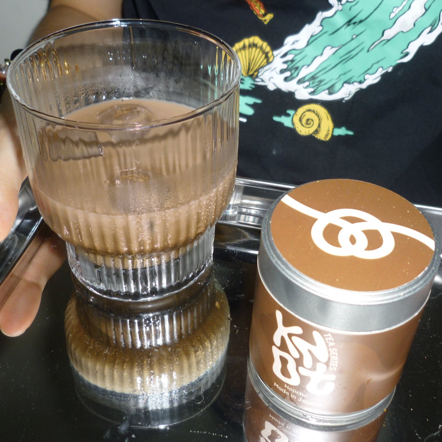 Glass filled with a hojicha latte next to a container of coffee on a mirror surface