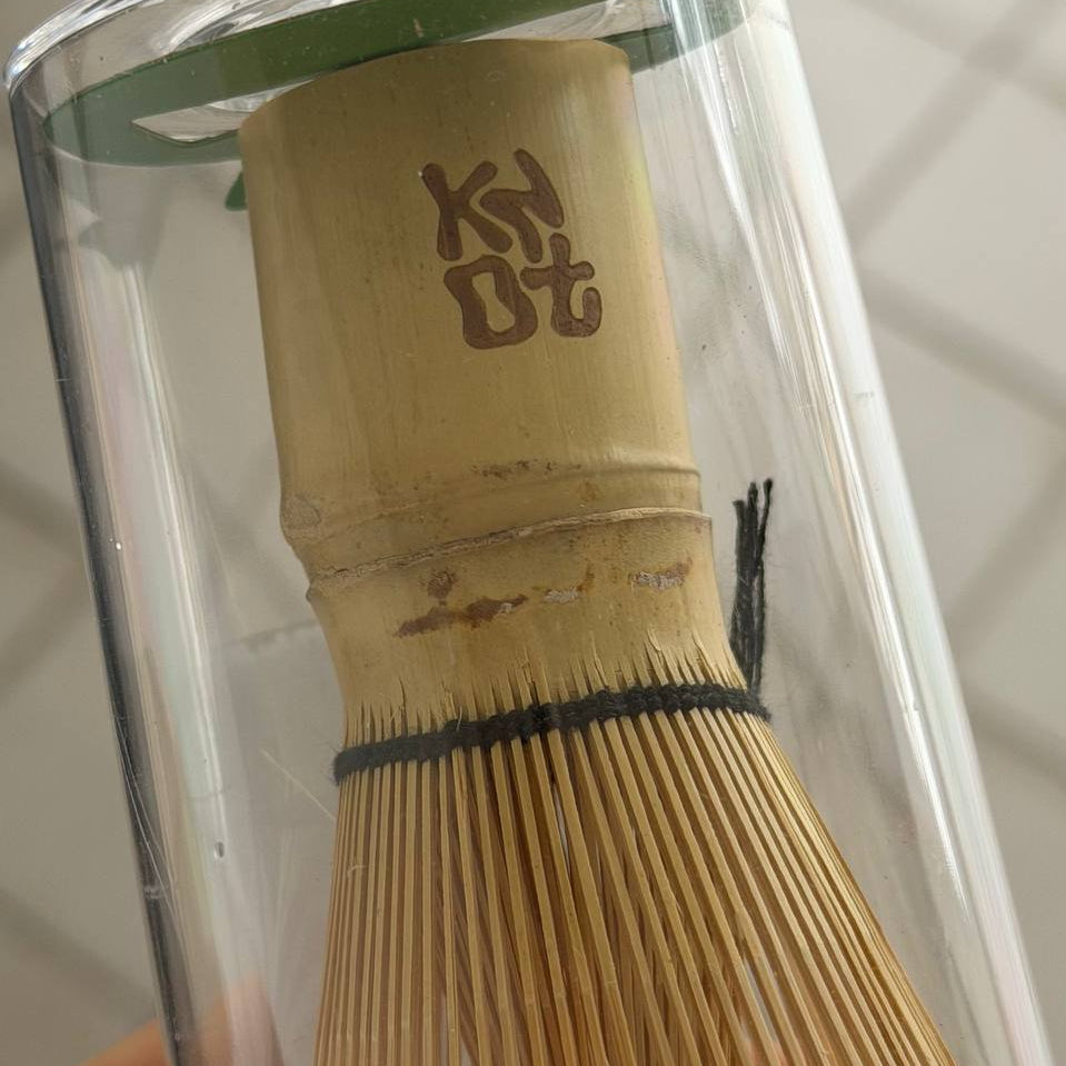 A close-up of a Japanese matcha whisk (chashaku) made of light tan bamboo, held inside a clear cylindrical glass container. The bamboo handle features a brown engraved character, and the whisk has a black rubber band around the bristles.